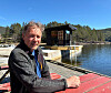 Mann sitter ved brygge foran en flytende badstuebygning på stille vann omgitt av skog.