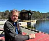 Mann sitter ved brygge foran en flytende badstuebygning på stille vann omgitt av skog.