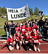 En gruppe elever i røde løpeklær står og sitter foran en stor «Heia Lunde»-plakat på en friidrettsbane.