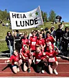 En gruppe elever i røde løpeklær står og sitter foran en stor «Heia Lunde»-plakat på en friidrettsbane.