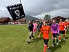 Fotballspillere og dommere går over en gressbane mens en svart klubbfane vaier i forgrunnen.