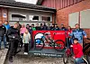 En gruppe mennesker står samlet rundt en rød sykkelvask med en sykkel foran en murbygning.